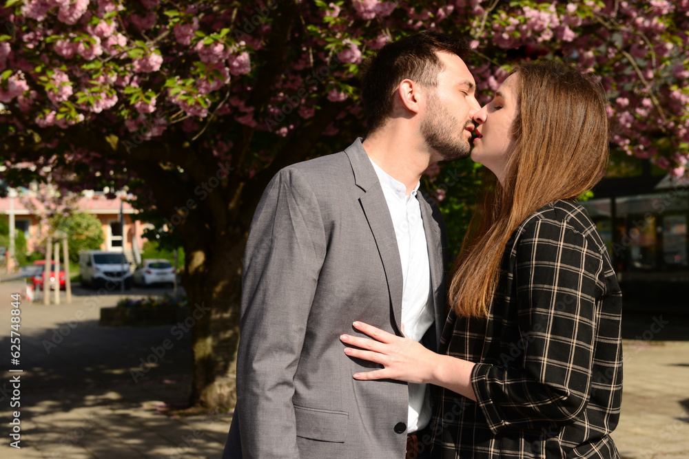 Beautiful loving young couple kissing near blooming tree on sunny spring day Beautiful loving young couple kissing near blooming tree on sunny spring day