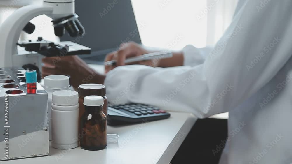 Medical worker in lab coat and sterile mask, doing a microscope analysis while her colleague are working behind Medical worker in lab coat and sterile mask, doing a microscope analysis while her colleague are working behind