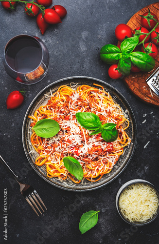 Spaghetti bolognese or pasta with minced meat in tomato sauce with green basil sprinkled with grated parmesan cheese and chianti red wine glass, dark table, top view