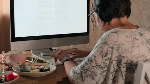 Adult female freelancer hunched over, sitting finishing urgent project, typing on keyboard in front monitor with white screen, plates of half-eaten Japanese rolls lying on desktop in home office. photo