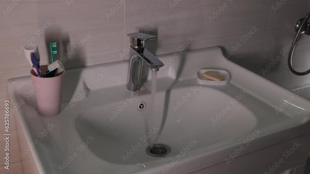 White washbasin from faucet dripping water tiled wall, hygiene items toothbrush and soap. Close-up side view of bathroom. Concept of amenities and comfort in modern life. White washbasin from faucet dripping water tiled wall, hygiene items toothbrush and soap. Close-up side view of bathroom. Concept of amenities and comfort in modern life.