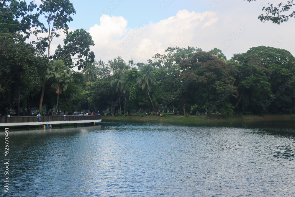 Beautiful central park view with blue sky and clouds. Lake and sky scenery. Scenic nature view inside a park with a big lake and green trees. Southeast Asian Park and beautiful natural scenery. Beautiful central park view with blue sky and clouds. Lake and sky scenery. Scenic nature view inside a park with a big lake and green trees. Southeast Asian Park and beautiful natural scenery.