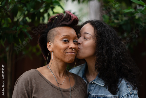 Two women express their love. One of them kisses the other one on the cheek. They are a lesbian couple, and Black and multiracial, expressing additional backstory.