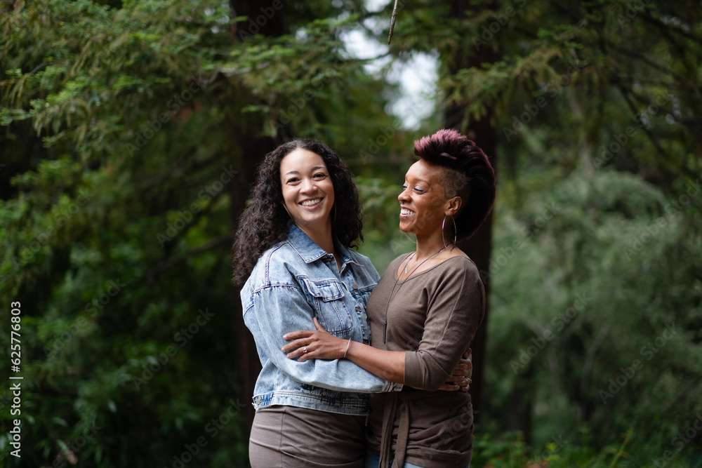 Naklejka premium With big smiles as they celebrate their engagement, a multiracial lesbian couple poses for portraits in a forest. They are facing each other and in a loving embrace. 