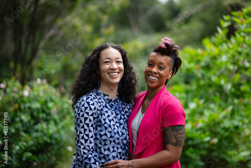A cheerful, joyful lesbian couple stands face to face in an affectionate embrace. Their happiness reflects how in love they are, celebrating their engagement. They are outdoors in a lush, green park.