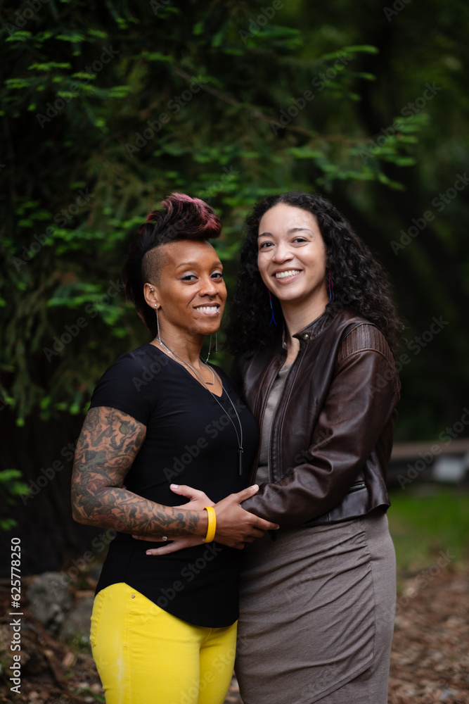 A young couple shares an embrace as they pose in a lush green park, surrounded by trees. Adding backstory, they are multiracial, lesbian, and self-identify as queer, with a slight edge to their style. A young couple shares an embrace as they pose in a lush green park, surrounded by trees. Adding backstory, they are multiracial, lesbian, and self-identify as queer, with a slight edge to their style.