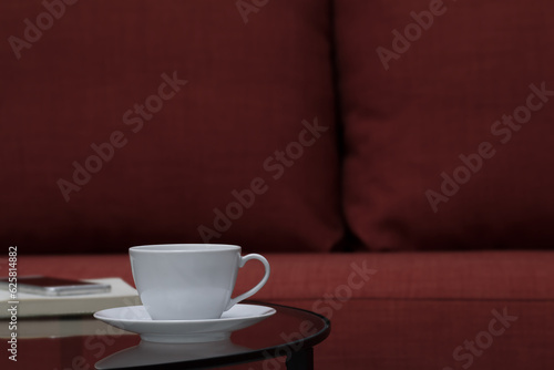 Clean white coffee cup and plate put on mirror table with blurred smart phone and notebook in the living room. Selective focus and copy space.