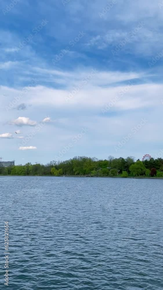 HD Video, vertical orientation -Scenic view from a tour boat on Herastrau Lake in the Park with the same name on a beautiful sunny day, with blue sky and white fluffy clouds, in Bucharest, Romania. HD Video, vertical orientation -Scenic view from a tour boat on Herastrau Lake in the Park with the same name on a beautiful sunny day, with blue sky and white fluffy clouds, in Bucharest, Romania.