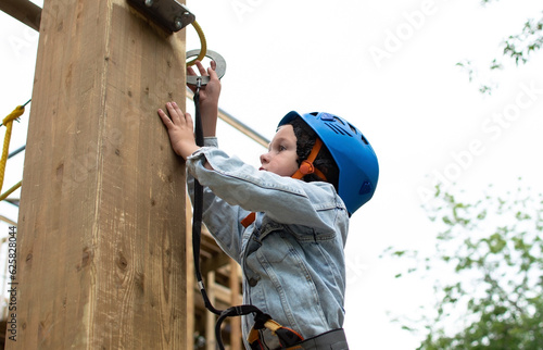 Happy child in helmet and protective gear enjoys classes in climbing adventure park on summer day. Kid is climbing on a rope playground