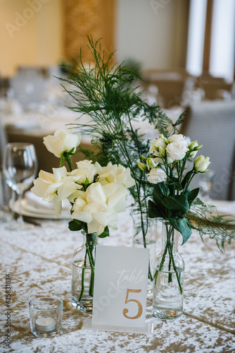 Table number 5. Stands, sign number five. Setup detail. Luxury reception. Elegant table decorated composition flowers in party area. Beautifully organized event. Wedding hall with atmospheric decor.