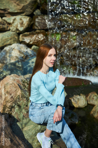 A stylish beautiful young girl is sitting on a rock by a waterfall. Photo shoot in the park