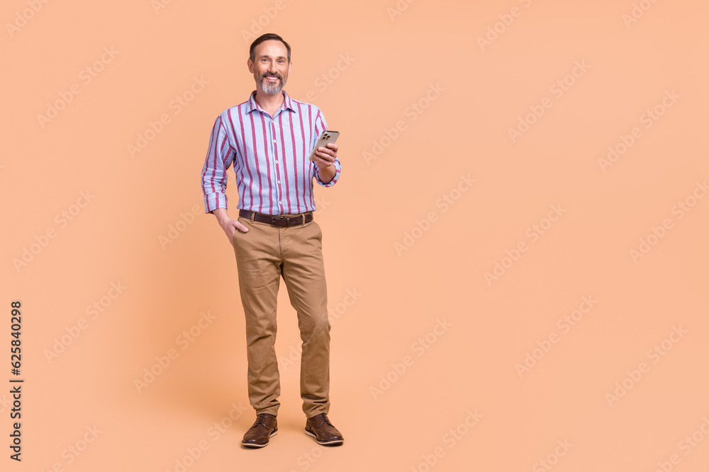Fototapeta premium Photo of positive cheerful confident man wear striped shirt brown trousers hold smartphone read post isolated on beige color background