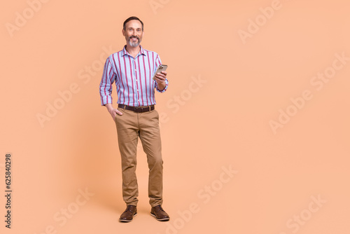 Photo of positive cheerful confident man wear striped shirt brown trousers hold smartphone read post isolated on beige color background