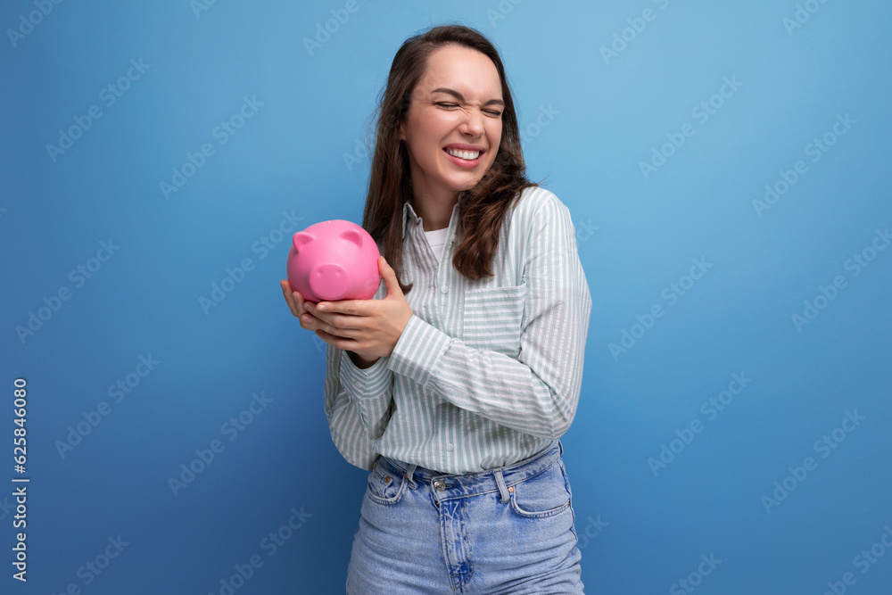 Joyful brunette young business woman in a shirt with a piggy bank on the background with copy pace Joyful brunette young business woman in a shirt with a piggy bank on the background with copy pace