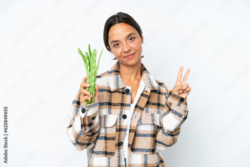 Young hispanic woman holding a green beans isolated on white background smiling and showing victory sign Young hispanic woman holding a green beans isolated on white background smiling and showing victory sign