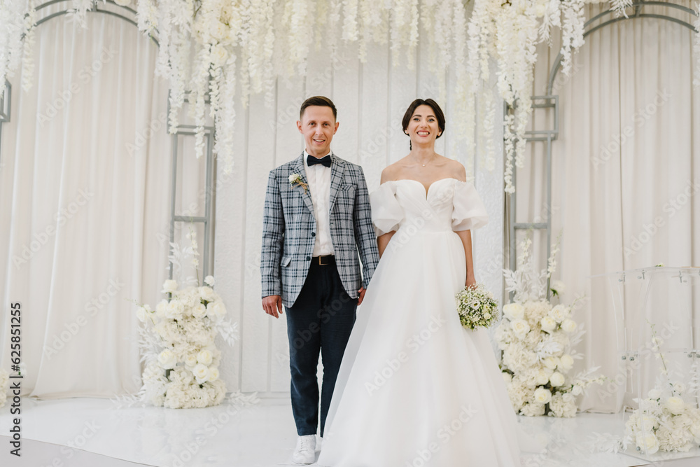 Bride and groom get married. Newlyweds with a bouquet, standing on wedding ceremony under arch decorated white flowers. Full length of attractive woman wearing dress and man while ceremony. Marriage. Bride and groom get married. Newlyweds with a bouquet, standing on wedding ceremony under arch decorated white flowers. Full length of attractive woman wearing dress and man while ceremony. Marriage.