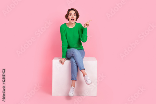 Full length photo of impressed lovely girl wear stylish clothes sitting podium arm direct empty space isolated on pink color background