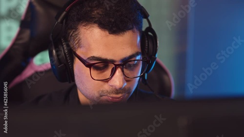 Chest up slowmo of concentrated Biracial guy wearing headset and eyeglasses playing pc game in dark esports club photo