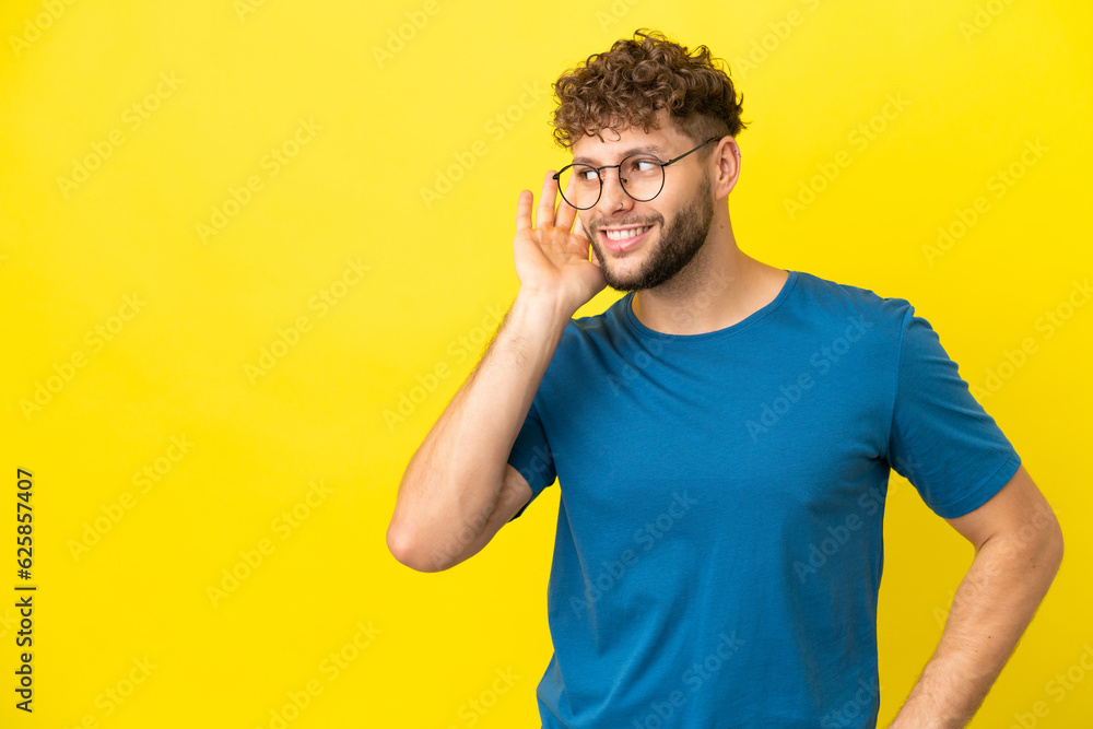 Fototapeta premium Young handsome caucasian man isolated on yellow background listening to something by putting hand on the ear