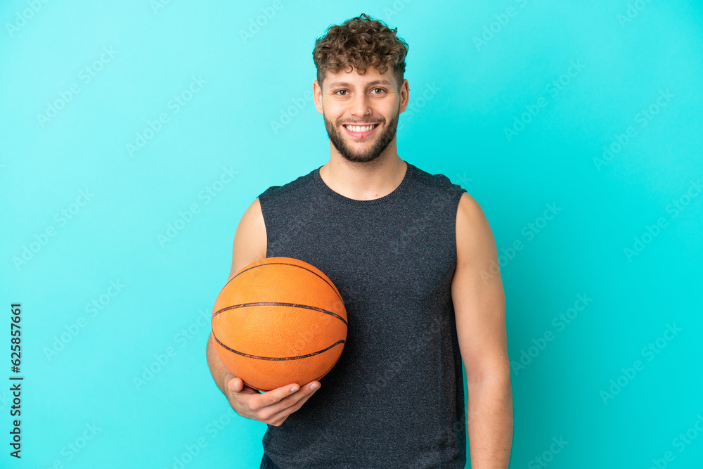 Handsome young man playing basketball isolated on blue background posing with arms at hip and smiling Handsome young man playing basketball isolated on blue background posing with arms at hip and smiling