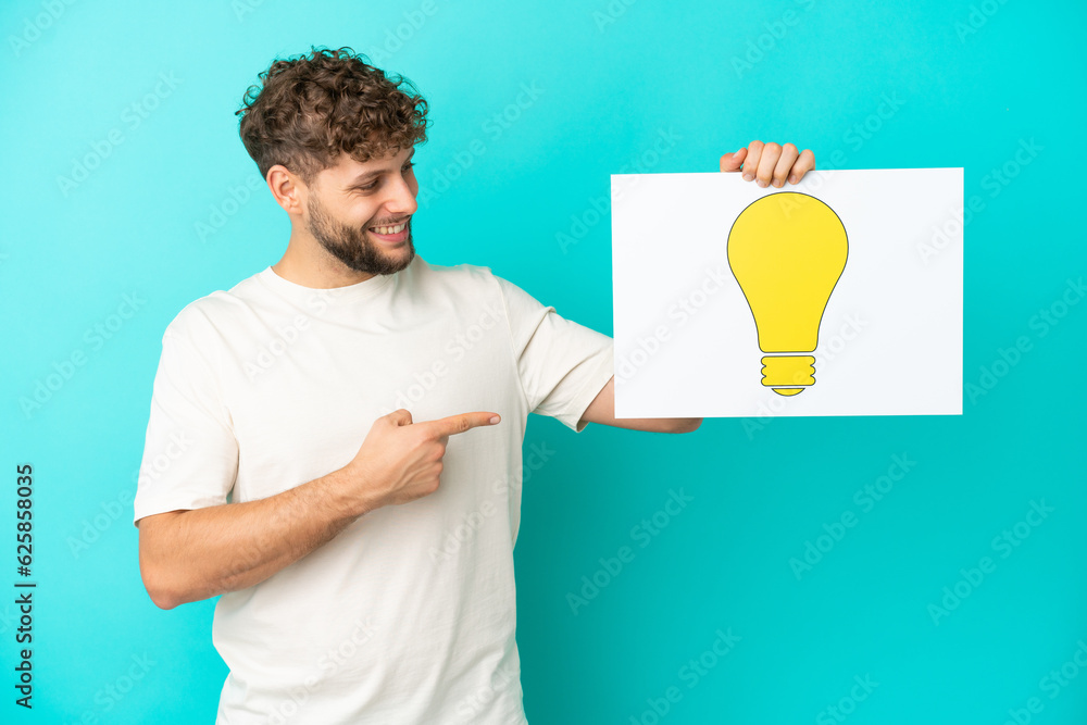 Young handsome caucasian man isolated on blue background holding a placard with bulb icon and pointing it Young handsome caucasian man isolated on blue background holding a placard with bulb icon and pointing it