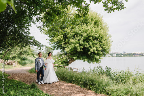 Bride and groom hold hands and walking in nature. Love story. Happy wedding day of marriage. Getting married outdoors. Newlyweds couple are walk near a big tree on the shore of a lake. © Serhii