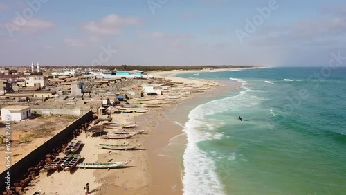 A sandy beach of turquoise waters in the fishing village of Kayar (Senegal). The aerial view advances as the waves reach the shore, which is peppered with a few colorful traditional fishing boats.