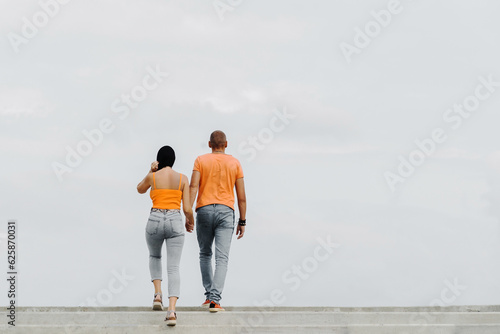 A young couple in orange t-shirts walk holding hands. The guy and the girl climb the stairs against the backdrop of a cloudy sky. Back view. Unrecognizable person. Space