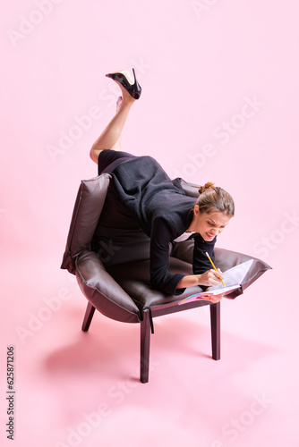 Anger and irritation. Business Woman lying on armchair in weird pose and putting notes on against pink studio background. Concept of business, working routine, deadlines, freelance, office, ad photo