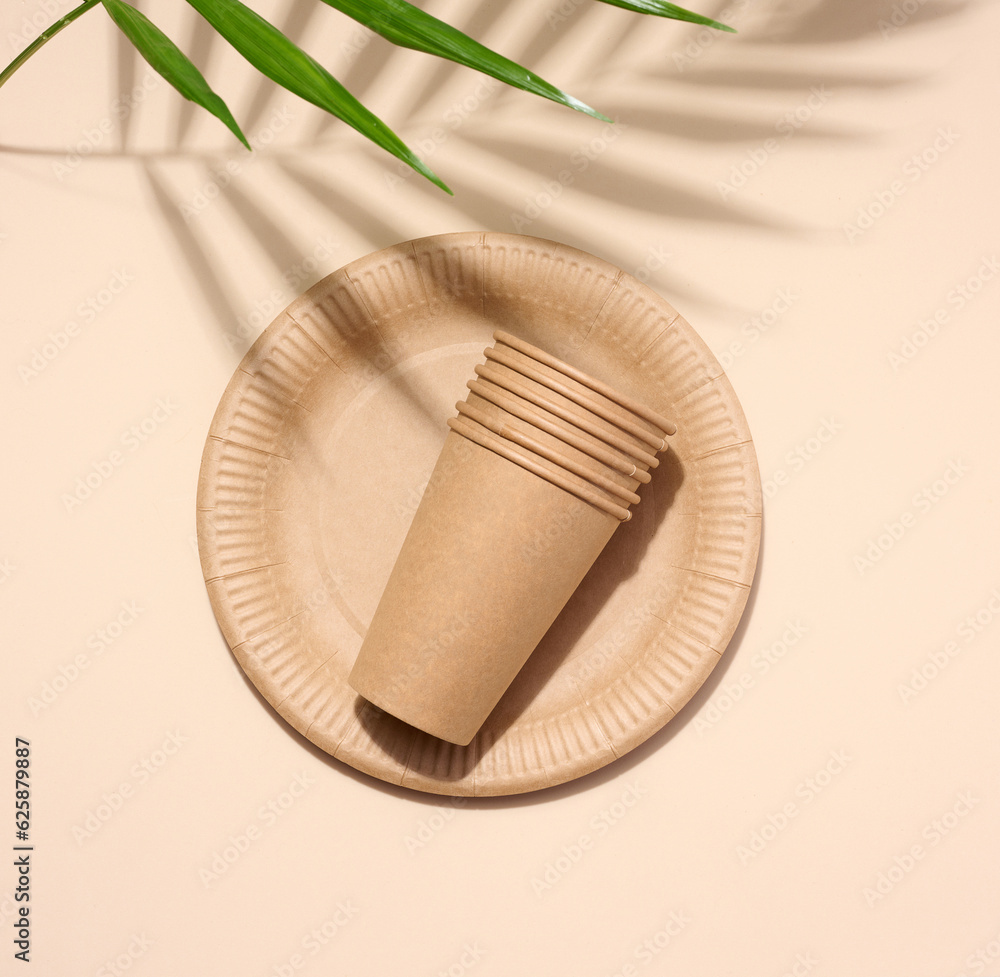 Brown paper plate, stack of paper cups, disposable tableware on a beige background, top view Brown paper plate, stack of paper cups, disposable tableware on a beige background, top view