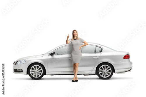 Full length portrait of a young woman leaning on a silver car and gesturing thumbs up
