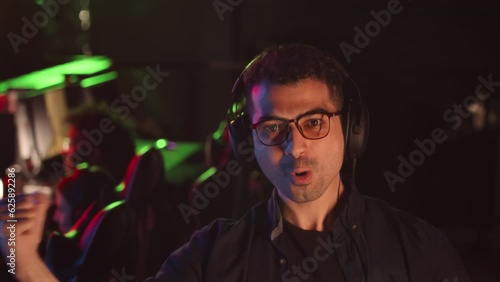 Slowmo waist up portrait of Middle Eastern gamer wearing headphones and eyeglasses rejoicing at camera in dark cybersport club photo