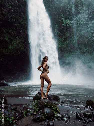 Beautiful woman in bikini posing near waterfall in tropical Bali. Traveler girl posing on river with powerfull waterfall