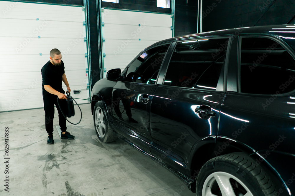 A male car wash employee applies car wash detergent to a black luxury car using a spray gun in the car wash box