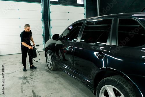 A male car wash employee applies car wash detergent to a black luxury car using a spray gun in the car wash box © Guys Who Shoot