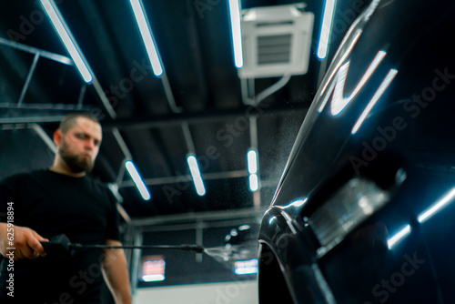 A male car wash employee applies car wash detergent to a black luxury car using a spray gun in the car wash box