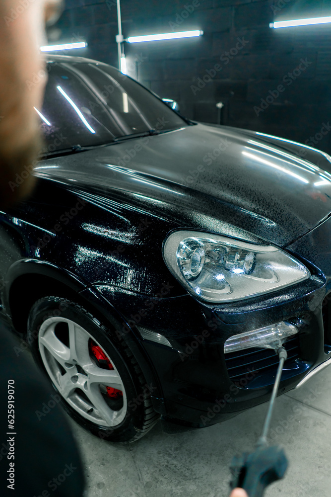 A male car wash employee applies car wash detergent to a black luxury car using a spray gun in the car wash box