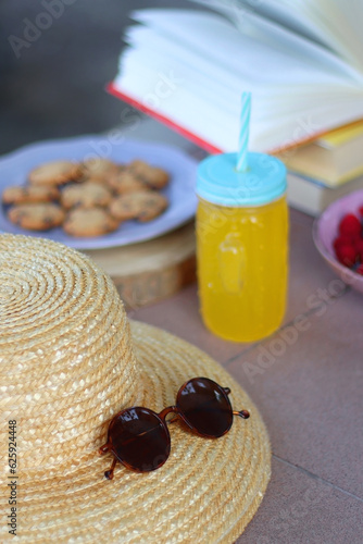 Straw hat, orange soda in the cactus shaped cup, bowl of fresh blueberreis and raspberries, plate of chocolate chip cookies, stack of books and sunglasses in the garden. Selective focus.