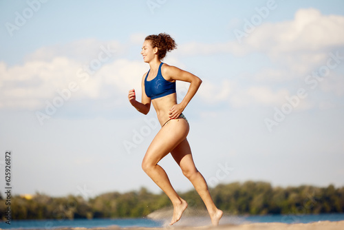 Young woman with slim, fit, healthy body running on beach on warm summer day outdoors. Concept of sport, active and healthy lifestyle, hobby, summertime, ad