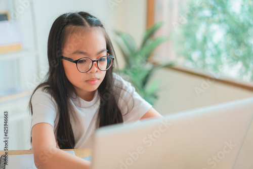 Asian school girl doing homework at laptop raising hand pointing finger . The girl does his homework. Home schooling. © kongga studio