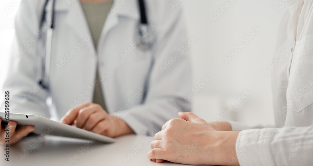Doctor and patient sitting at the desk in clinic office. The focus is on female physician's hands using tablet computer, close up. Perfect medical service and medicine concept Doctor and patient sitting at the desk in clinic office. The focus is on female physician's hands using tablet computer, close up. Perfect medical service and medicine concept