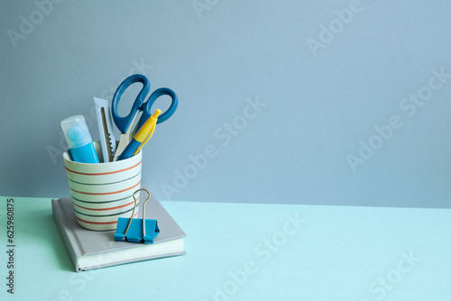 Obraz na płótnie Writing supplies in pen holder and notebook on blue desk. blue wall background. workspace w salonie