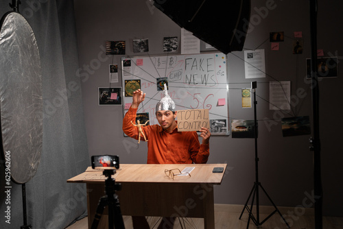 Strange young man in tinfoil hat recording video or live streaming using phone in home studio. Fake news conspiracy theories