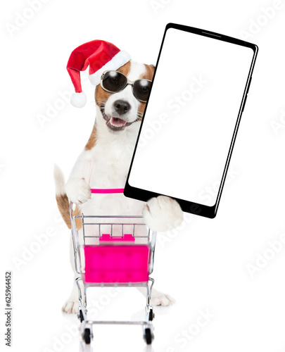 Jack russell terrier puppy wearing sunglasses and red santas hat pushing a shopping cart and shows big smartphone with white blank screen in it paw. isolated on white background © Ermolaev Alexandr