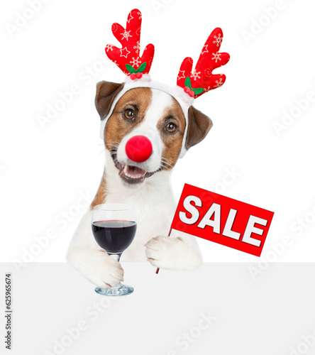 Jack russell terrier puppy dressed like santa claus reindeer  Rudolf holds glass of red wine above empty white banner and shows signboard with labeled "sale". isolated on white background © Ermolaev Alexandr
