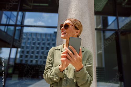 Stylish woman entrepreneur in sunglasses using phone whiile standing on modern building background  photo