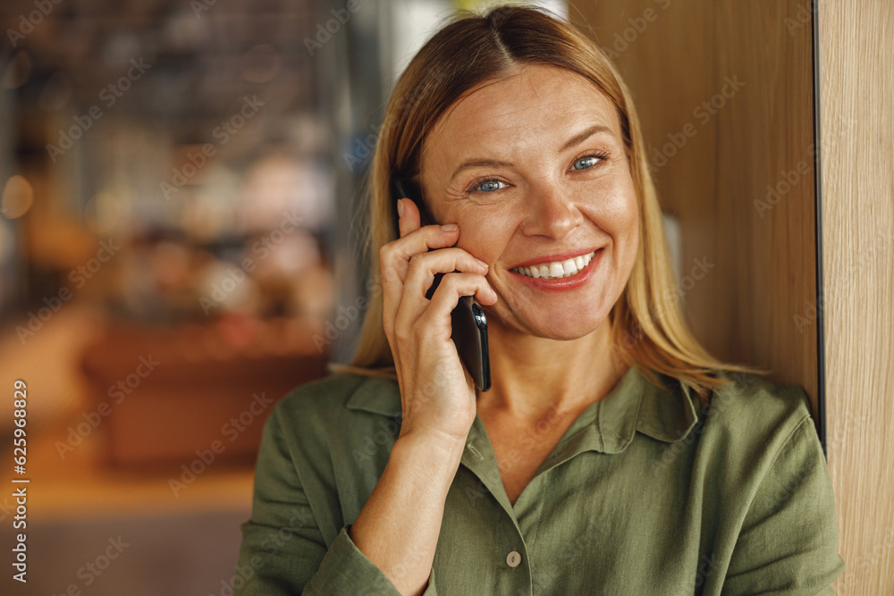 Smiling businesswoman is talking phone with client while standing near wooden wall in cafe Smiling businesswoman is talking phone with client while standing near wooden wall in cafe