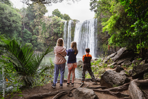 Three people, a mum and two children at an idyllic location looking at a waterfall, with their backs turned. This was taken at Whangarei Falls, New Zealand.