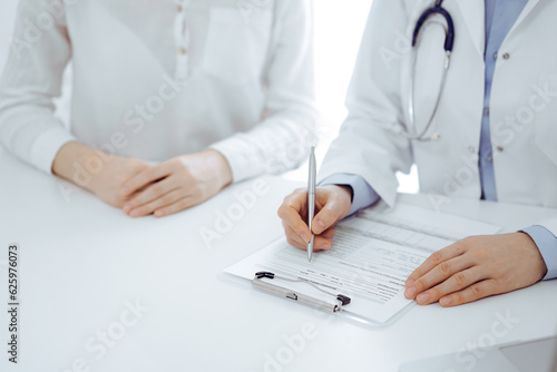 Doctor and patient sitting near of each other at the desk in clinic. The focus is on female physician's hands filling up the medication history record form or checklist, close up. Medicine concept