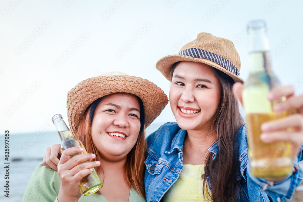 Two Asian women partying at evening beach holding beer bottles smiling happy having fun together. Celebration concept.